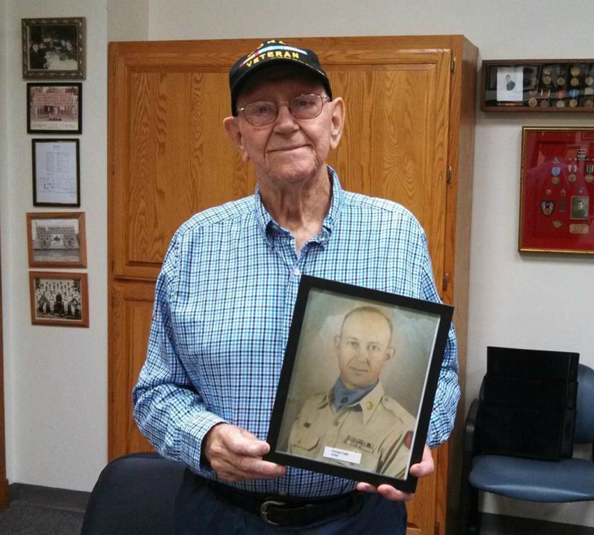 Man in blue plaid shirt and Korea Veteran hat holds up framed picture, and smiles into camera